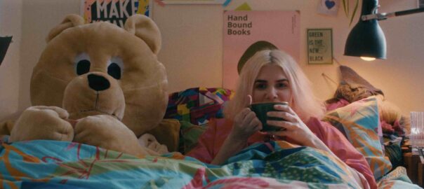A giant teddy bear sits in bed beside a blonde woman on her phone, tucked under a colorful blanket.